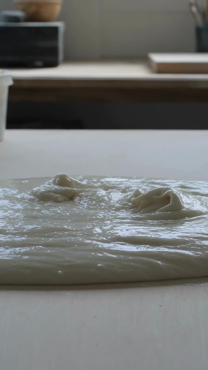 Vertical video: Pouring from above, wet batter forming smooth sheet on kitchen bench with tub