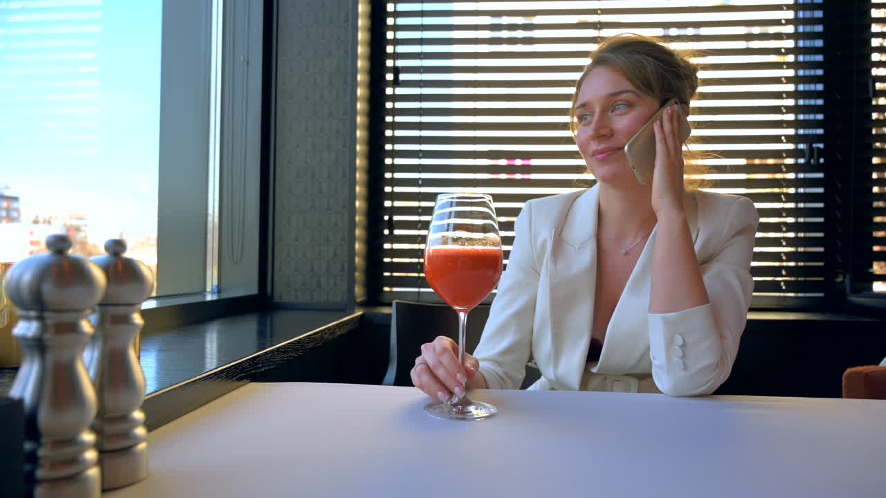 Woman in white blazer talking on the phone with a carrot juice on the table at a restaurant