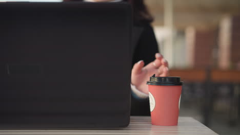 close-up back view of laptop on table, person in black coat sitting with coffee cup dropping it on table in restaurant setting, blurred background