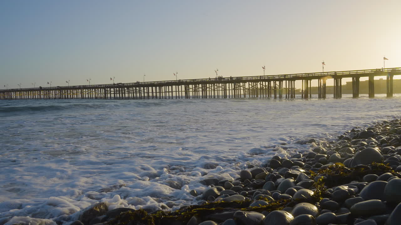 캘리포니아 남부에 위치한 벤츄라 해변의 기슭을 따라 파도가 부서지는 동안 백그라운드에서 일몰과 함께 벤츄라 부두의 패닝 샷