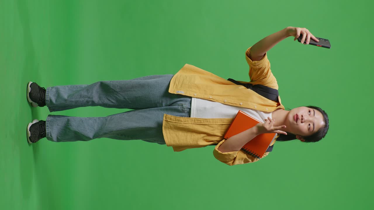 Full Body Of Asian Woman Student With A Backpack And Some Books Taking Photo On Smartphone While Standing In The Green Screen Background Studio