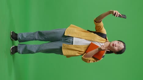 full body of asian woman student with a backpack and some books taking photo on smartphone while standing in the green screen background studio