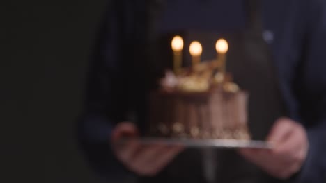 studio shot of person carrying decorated chocolate birthday celebration cake with lit candles against black background