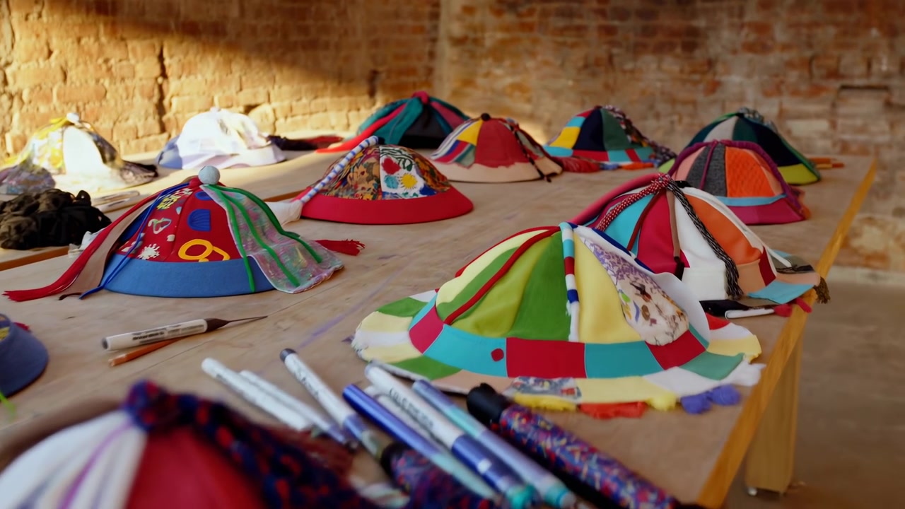 Calda vista all'altezza degli occhi che mostra file di cappelli patchwork colorati fatti a mano su un tavolo di legno all'interno di un laboratorio ristrutturato, con la luce del sole che rivela le texture dei tessuti, le finiture intrecciate, le nappe, le cuciture e.