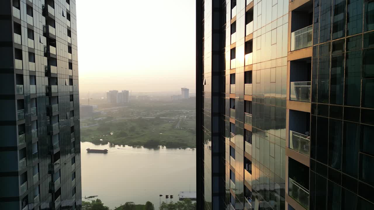 Toma cinematográfica con dron moviéndose entre rascacielos de cristal revelando el río Saigón y los humedales al amanecer, paneando a la izquierda hacia una vasta vista del río con barcos de carga y un puente en la neblina dorada de la mañana