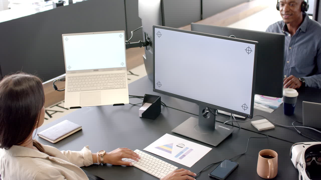 Working in modern office, woman using laptop and monitor, African American man smiling nearby, copy