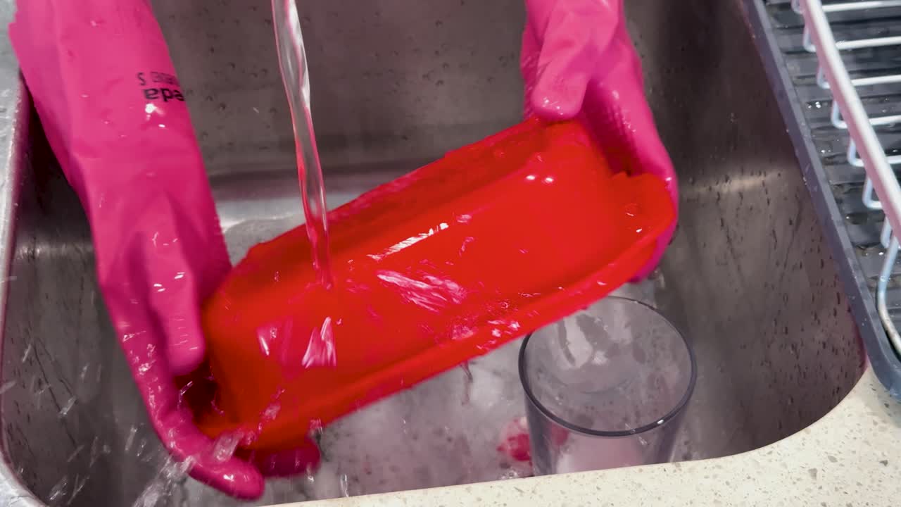 Person wearing pink gloves thoroughly washes a red rectangular dish under running water in a stainless steel kitchen sink, with bright, even lighting and steady overhead camera angle