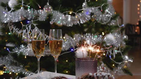 two glasses of sparkling champagne next to the burning sparklers in a candlestick on a table against the background of a decorated christmas tree