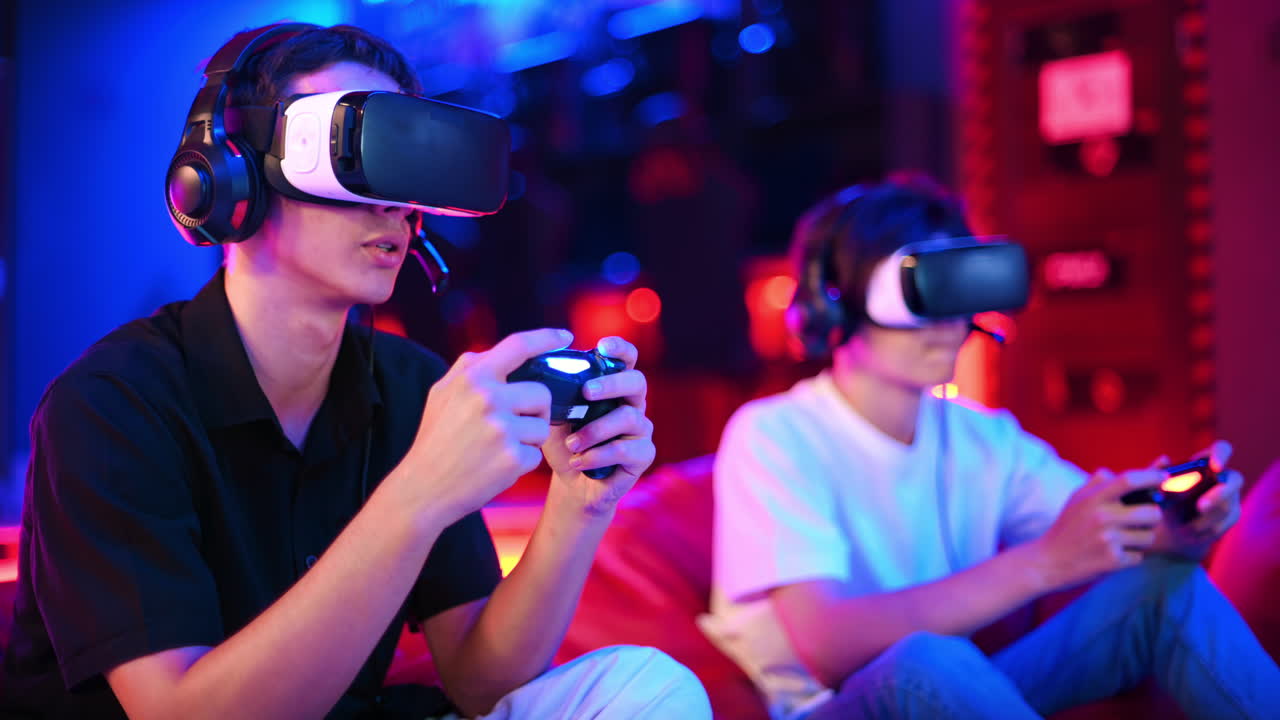 Two teen friends are playing a game console in VR headset and headphones using gamepads, talking and smiling while sitting on bean bags. Blue and red illumination