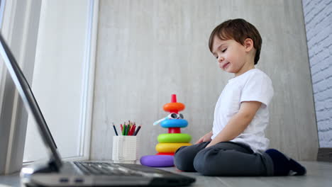 adorable child in white t-shirt and grey pants sits on his knees on the floor. lovely baby pressing keys on the keyboard. low angle view.