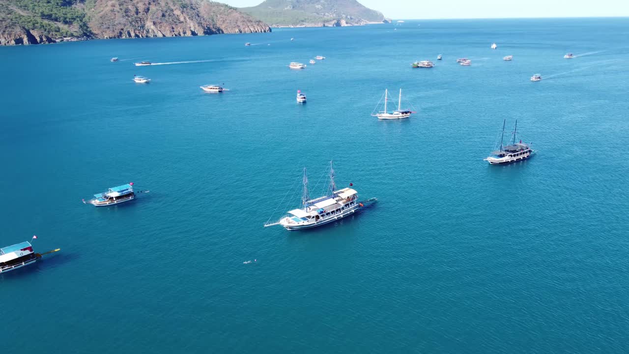 Aerial drone view showing scattered sailboats across wide blue expanse of Gokova Bay with coastal hills and calm summer conditions in Mugla Province under bright daylight