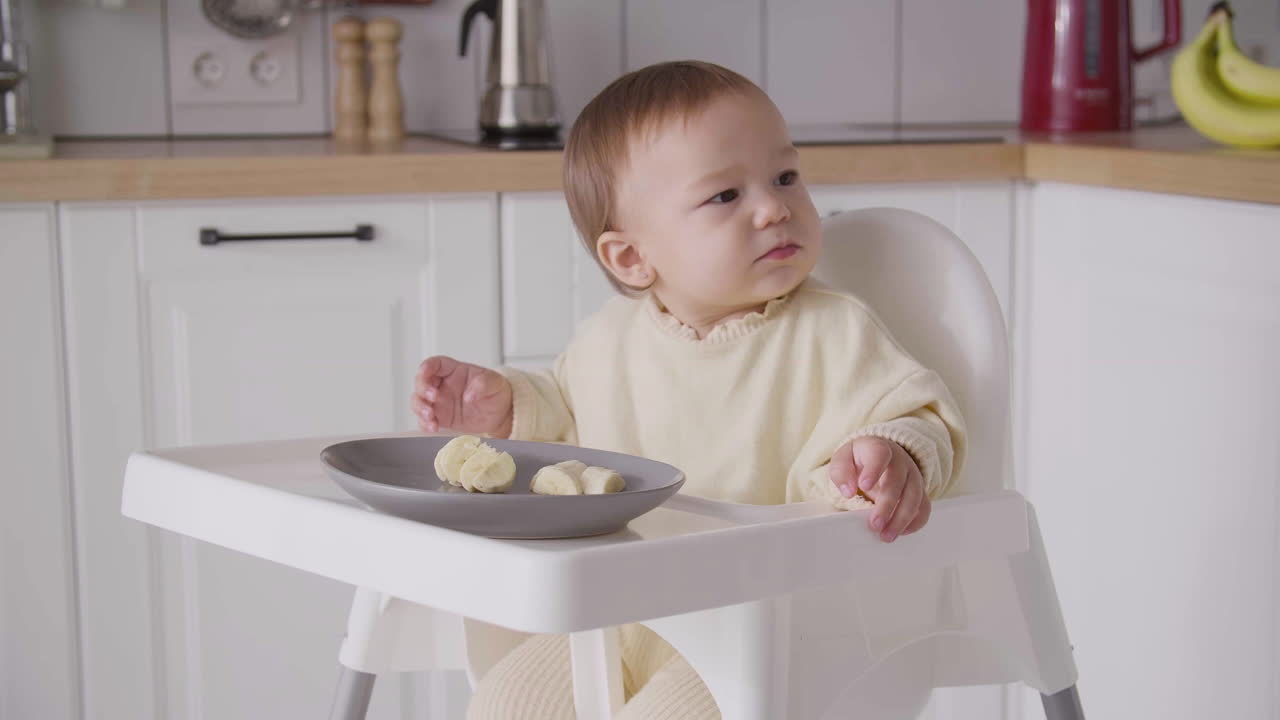 linda niña comiendo rodajas de plátano sentada en su silla alta en la cocina