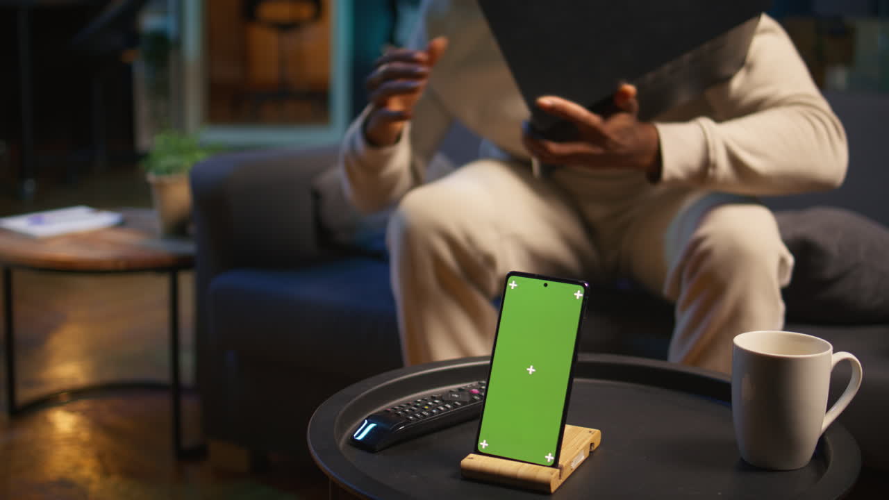 trabajador masculino negro remoto acostado en el sofá al lado de la pantalla de llave de croma, disfrutando del ocio y el tiempo libre para la relajación en su moderno apartamento de loft con luces de neón y una televisión inteligente. cámara b.