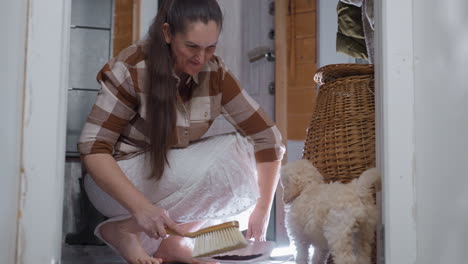 pet owner sweeping spilled soil near door while playful puppy explores indoors, using brush to collect debris on tile floor by brick wall under lamp light, capturing cleanup moment in cozy home entry