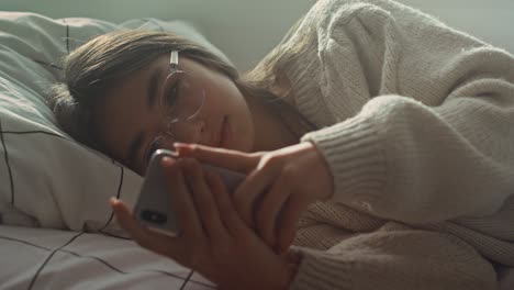 Close-up-of-caucasian-teenage-girl-browsing-phone-while-lying-on-side-in-bed