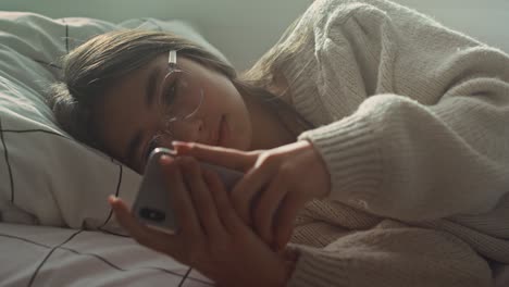 primer plano de una adolescente caucásica navegando por el teléfono mientras está acostada de lado en la cama