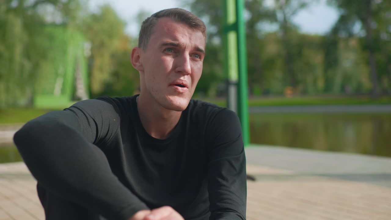 white man cooling down after workout, sitting on wooden dock by pond, wiping sweat from forehead, catching breath, contemplative gaze, black longsleeve training top, green park background, soft