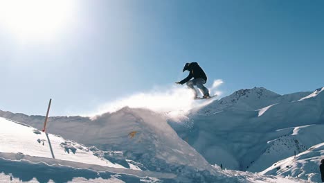 slow motion, drone shot, wide: pro snowboarder jumping in fresh snow, spraying snowflakes over sun. male freerider takes off in the air on his snowboard on sunny day on the mountain slopes