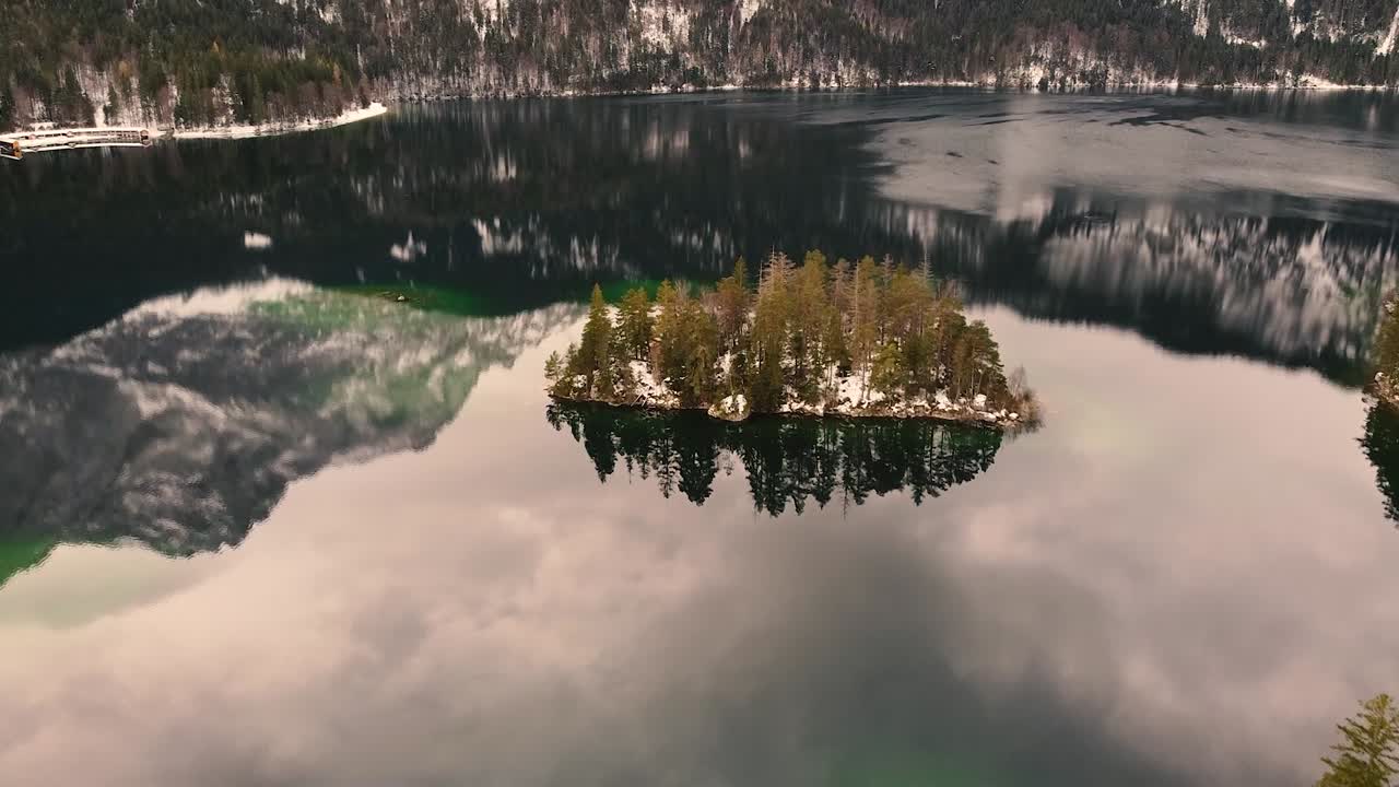 :
Aerial view of tiny island covered with pine trees in middle of clear lake reflecting surrounding snowy mountains and sky, creating peaceful and cinematic winter landscape in Iceland
