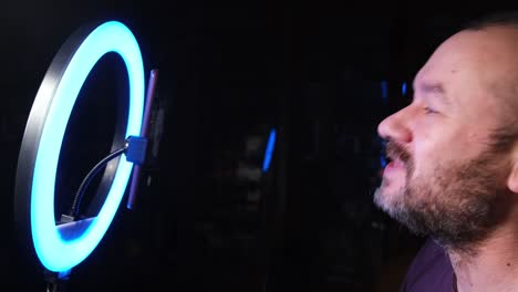 an unshaven man sits in front of a ring lamp and a mobile phone, vlogging.