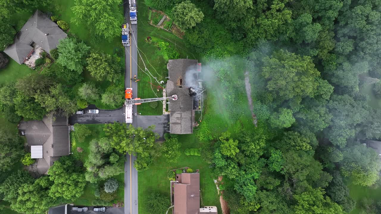 Fire department with ladder extinguishing burning house with toxic fumes in American suburb. Several hose and spraying water.Aerial top down shot