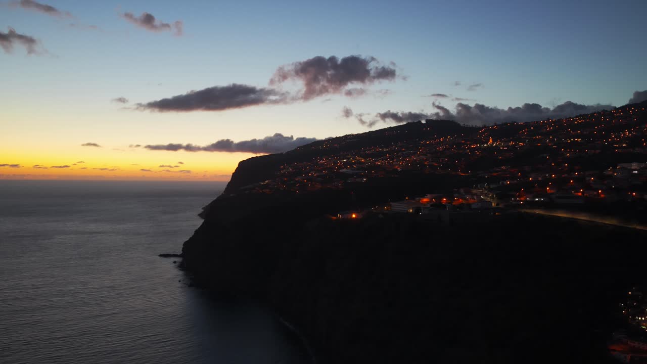 Calheta village on cliffside at sunset, illuminated town, coastline, and beautiful sky, Madeira, Portugal. Aerial drone lateral view