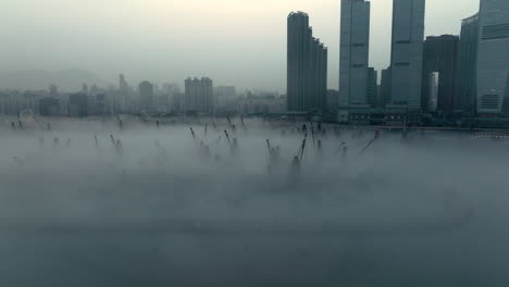 Suspenseful-aerial-shot-of-loading-barge-cranes-poking-out-of-coastal-morning-fog-with-urban-high-rise-silhouette-in-the-background