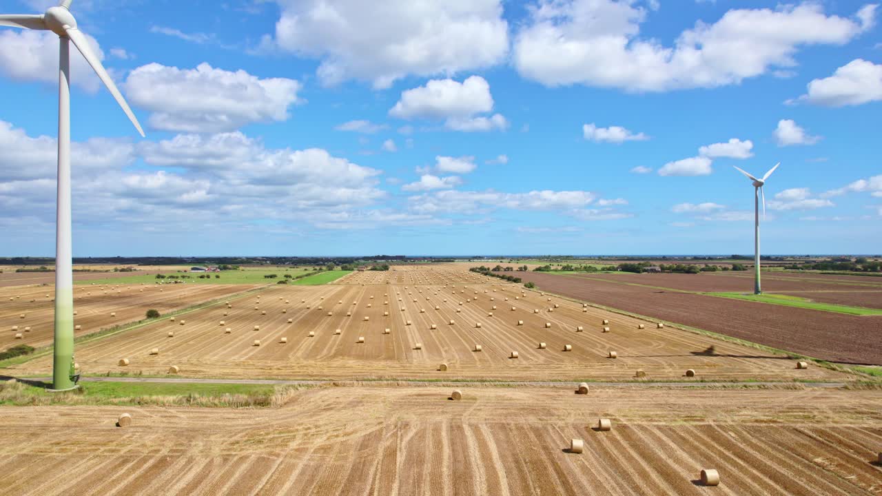 Aerial video footage immerses us in the tranquility of wind turbines in a row, gently spinning amidst a Lincolnshire farmer's freshly harvested field, framed by golden hay bales
