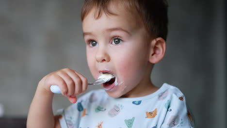 cute face of a lovely toddler smudged with dairy. sweet baby boy eating yoghurt with spoon. close up.