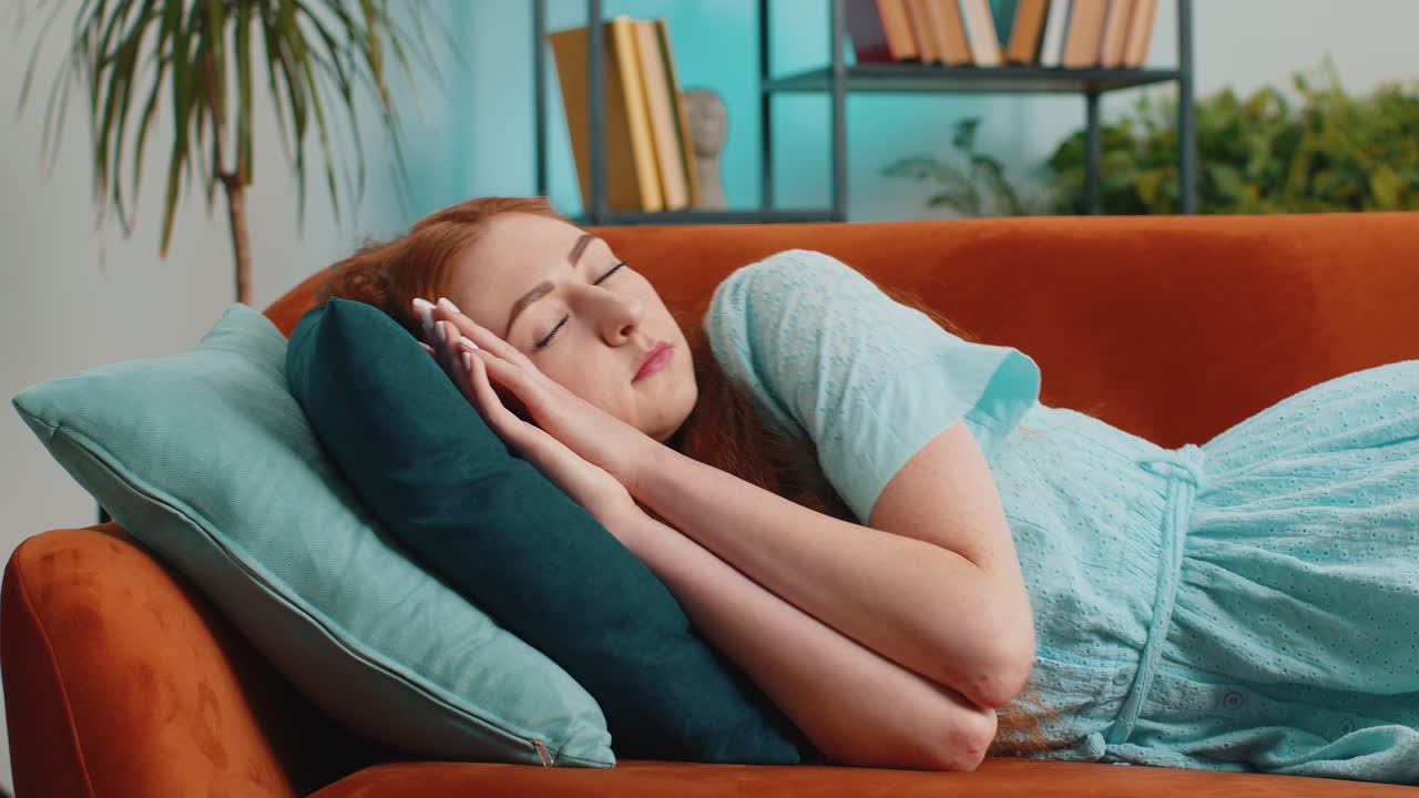Tired young redhead woman lying down in bed taking a rest at home napping falling asleep on sofa