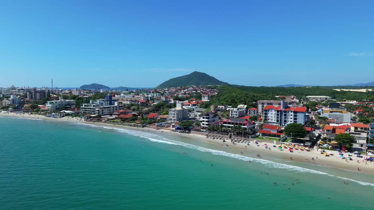 Praia dos Ingleses na Ilha de Santa Catarina durante a alta temporada de verão, com turistas ao longo do litoral atlântico sob as colinas da Mata Atlântica da Serra do Mar, em voo suave de drone.