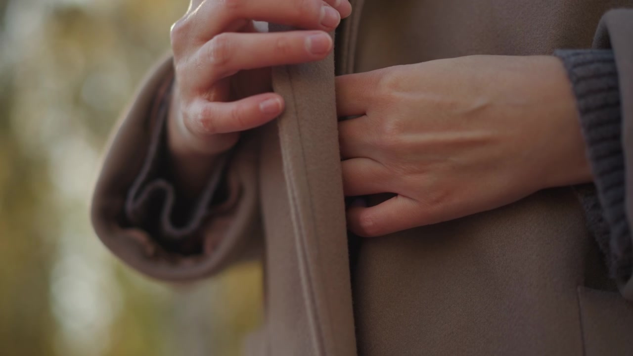 Primo piano di una donna che sistema la cintura e i dettagli del tessuto sul suo cappotto marrone, concentrandosi sui movimenti delle mani e sulla texture. Una luce soffusa evidenzia le proprietà del materiale mentre la donna manipola il capo