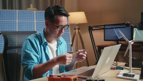 asian man looks at wind turbine while working with laptop next to the solar cell at the office