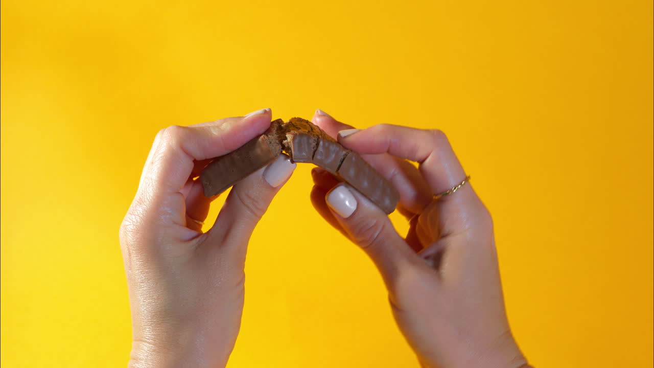 Close-up of two hands with manicured nails breaking a chocolate-coated protein bar in half, revealing the dense texture of the filling against a vibrant yellow backdrop for a rich satisfying visual