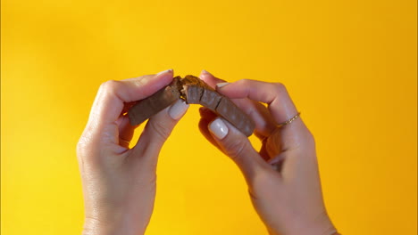close-up of two hands with manicured nails breaking a chocolate-coated protein bar in half, revealing the dense texture of the filling against a vibrant yellow backdrop for a rich satisfying visual