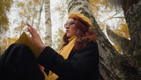 woman in yellow beret and scarf seated alone, leaning against tree, carefully opening yellow-covered book, surrounded by golden autumn leaves and birch trees, she enjoys quiet reading and reflection