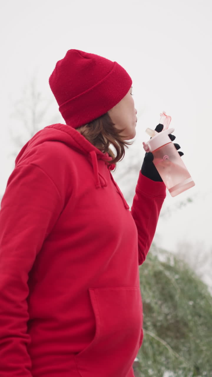 lady in red beanie and hoodie sipping water from pink bottle outdoors, winter park setting, snow-covered trees, benches in blurred background, fresh air, cold weather, peaceful outdoor moment