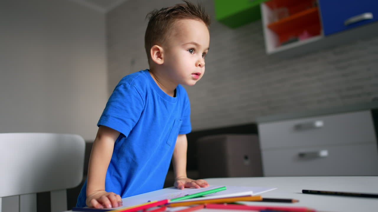 Un encantador niño pequeño con camiseta azul está de pie en su escritorio con lápices. El niño mira al frente y habla.