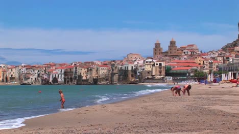 small waves break near houses along a shoreline as children play in the sand in cefalu italy