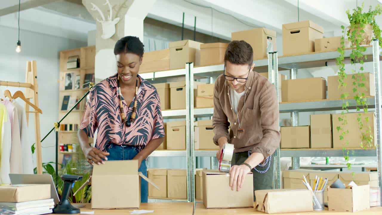 African American young woman designer and caucasian designer man wrapping a parcel and closing box with sticky tape while preparing order in fashion clothing shop