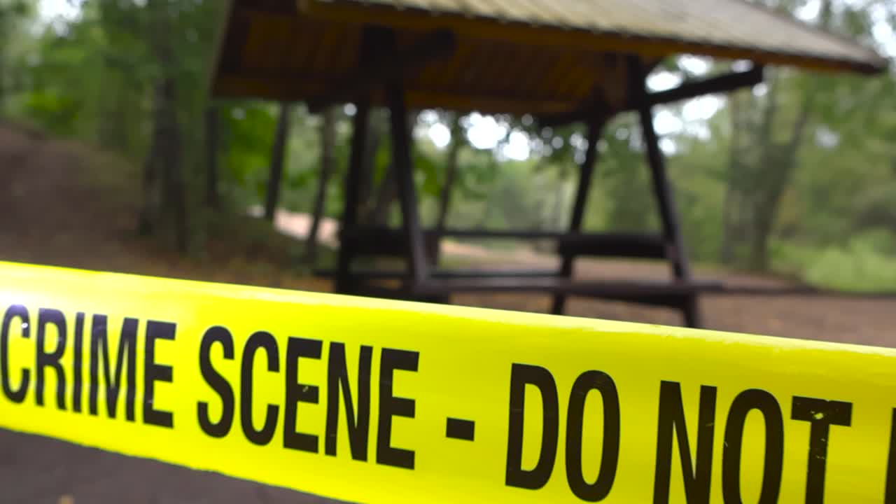 Closeup tracking moves along yellow crime scene tape stretched across forest park camping ang grilling area. Police tape in focus, campground with wooden picnic table in bokeh on moody, overcast day