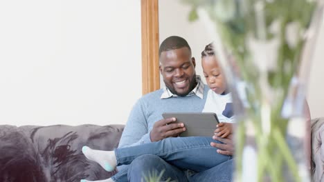 Happy-african-american-father-sitting-on-sofa-with-son-on-lap-using-tablet,-in-slow-motion