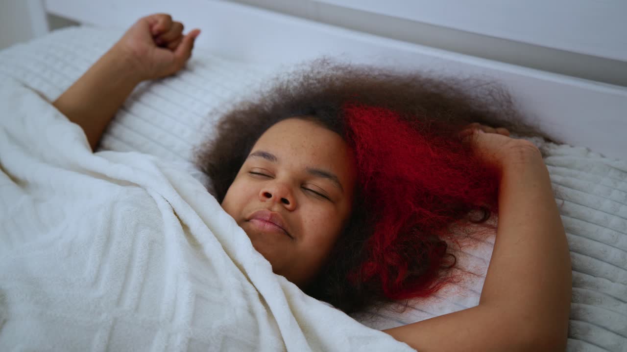 Young Black woman with vibrant red dyed hair leisurely stretching and smiling while waking up in cozy bedroom, embodying peaceful morning relaxation and wellness