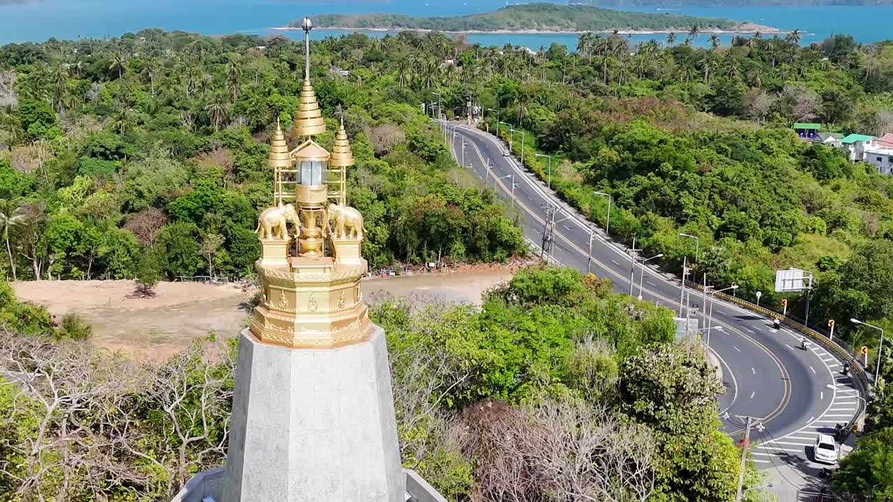 A golden spire stands tall above a winding coastal road surrounded by lush greenery and ocean views.