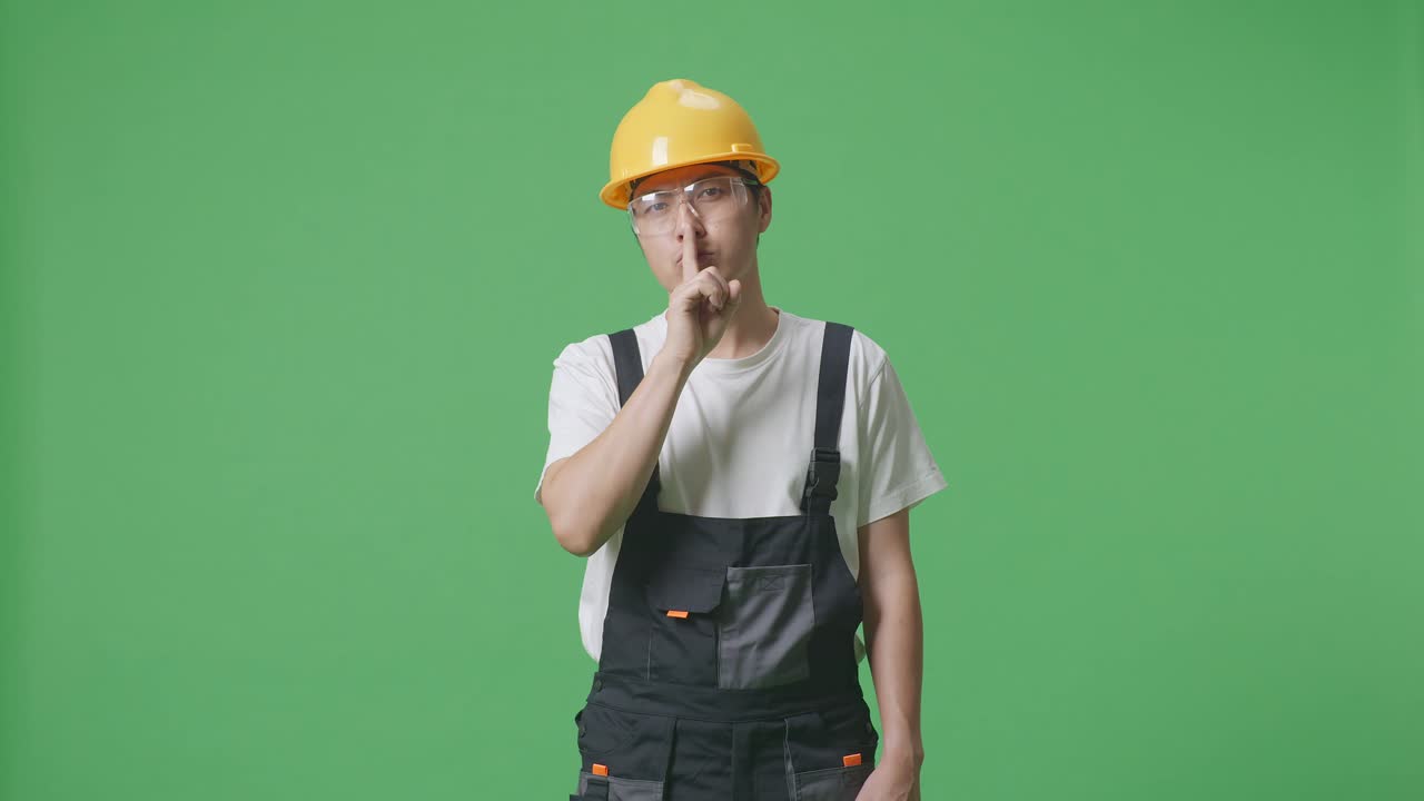 Asian Man Worker Wearing Goggles And Safety Helmet Looking At Camera And Making Shh Gesture While Standing In The Green Screen Background Studio