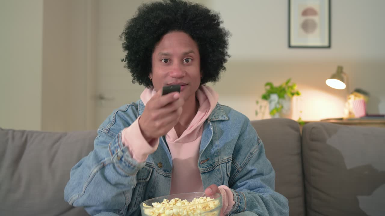 Afroamerikansk mand i midaldrende alder, der trykker på fjernbetjeningen, reagerer på programmet og spiser popcorn i stuen