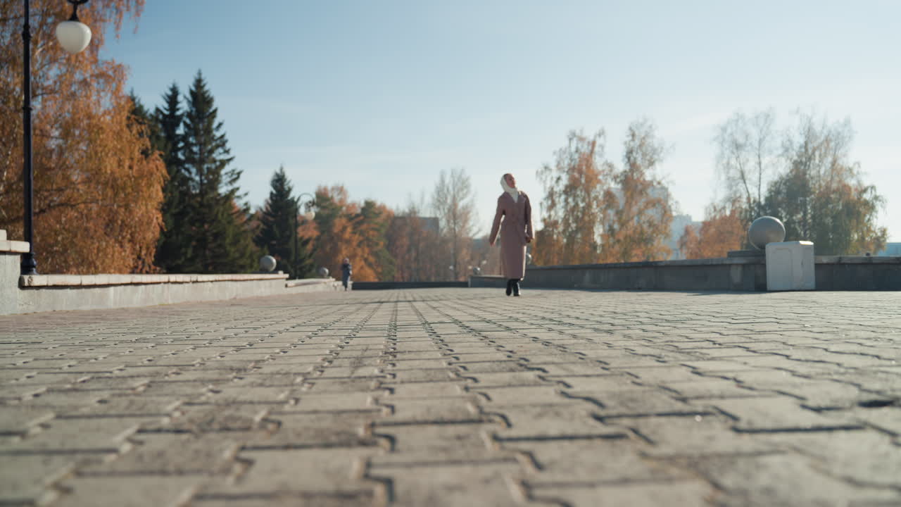 Asian woman walking sunlit promenade wearing hijab and coat, approaching camera across cobblestone pavement with lamppost and autumn trees in background, gentle smile and confident stride, closeup