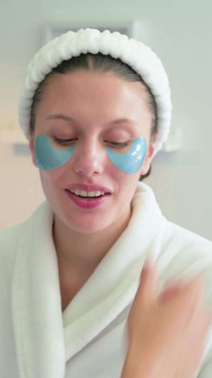 Vertical video: Hands placing blue eye pads on woman at home for care, she using jar, wearing robe