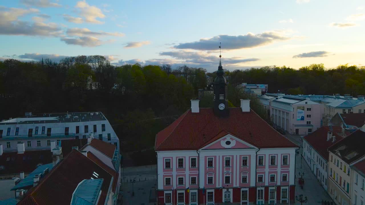 Aerial drone footage flying backwards reversing and revealing a dark black Tartu town hall clocktower during sunset golden hour in Tartu central city that has cobblestone roads and old historic houses