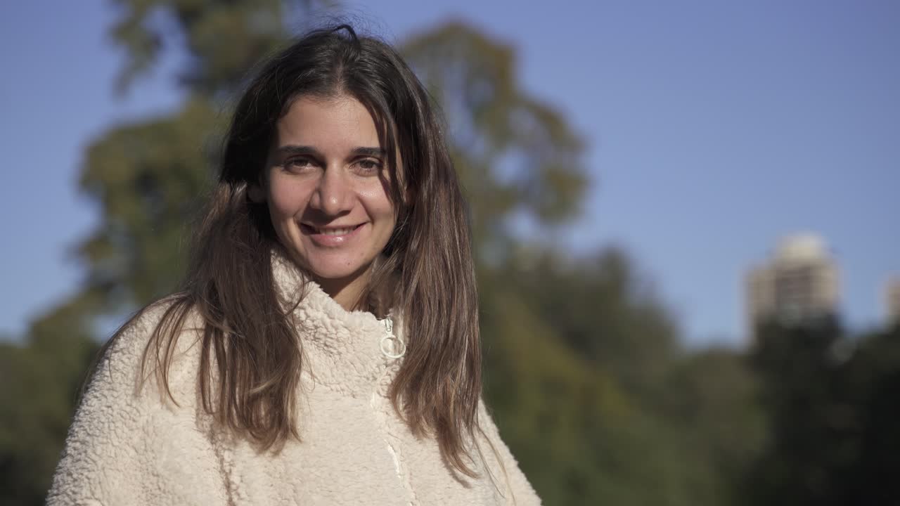 retrato de 4k de una joven encantadora mirando a la cámara y sonriendo iluminada por el sol contra el cielo azul y los árboles en el fondo, de cerca